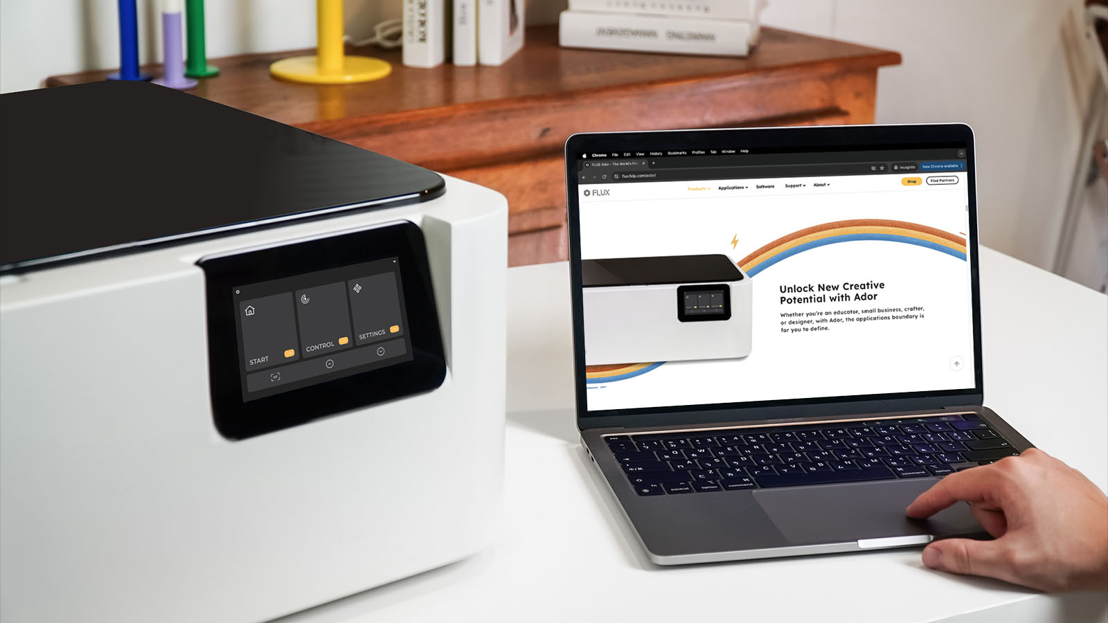 Ador laser cutter on a desk next to a laptop displaying the Ador product page.