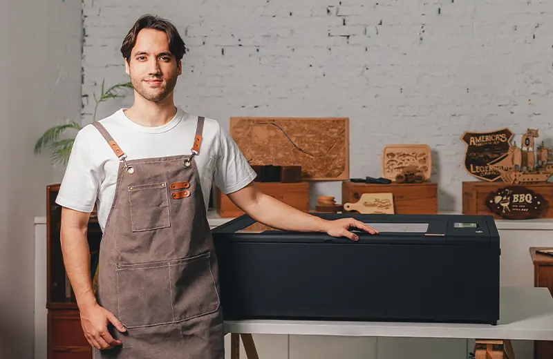 A man in a work apron standing next to a laser cutter, with wooden engraved signs and boxes on the shelf behind him.